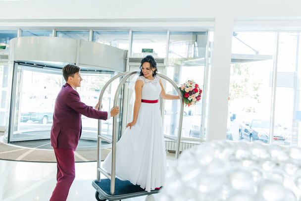 Ο γαμπρός σε ένα μπορντό κοστούμι και η νύφη σε ένα λευκό φόρεμα με μια κόκκινη κορδέλα και ένα μπορντό μπουκέτο, οι νεόνυμφοι σε μια φωτογράφηση, μια βόλτα στο γάμο - Φωτογραφία, εικόνα