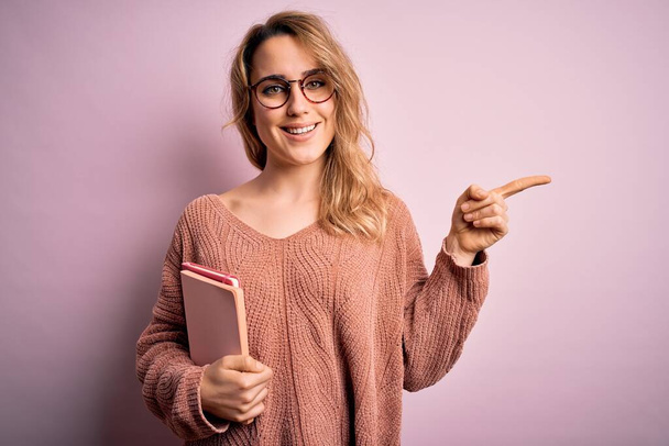 Mladá krásná blondýna žena nosí brýle drží notebook přes růžové pozadí velmi šťastný ukazující s rukou a prstem na stranu - Fotografie, Obrázek