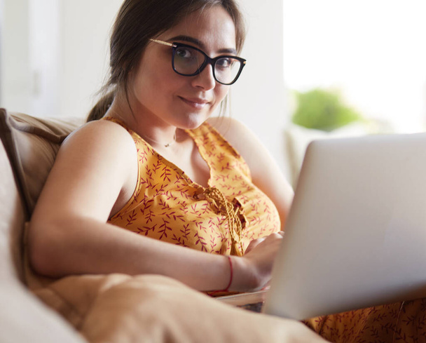 Young woman sitting on couch working on laptop computer at home - 写真・画像