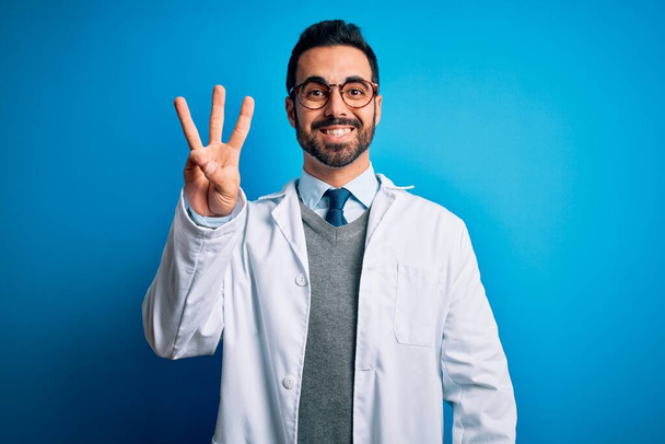Young handsome doctor man with beard wearing coat and glasses over blue background showing and pointing up with fingers number three while smiling confident and happy. - Photo, Image