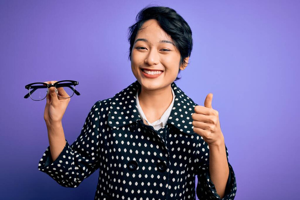 Mladý krásný asijské optický dívka držení vidění brýle přes izolované fialové pozadí šťastný s velkým úsměvem dělá ok znamení, palec nahoru s prsty, vynikající znamení - Fotografie, Obrázek