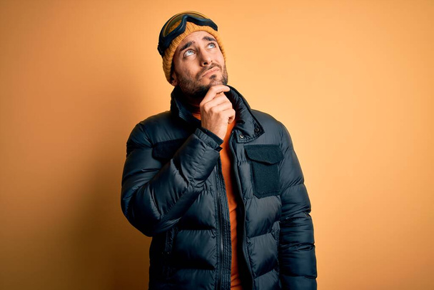 Young handsome skier man with beard wearing snow sportswear and ski goggles with hand on chin thinking about question, pensive expression. Smiling with thoughtful face. Doubt concept. - Fotoğraf, Görsel