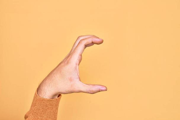 Hand eines kaukasischen jungen Mannes mit Fingern über isoliertem, gelbem Hintergrund, der unsichtbare Dinge pflückt und nimmt, Gegenstand mit Fingern hält, die Raum zeigen - Foto, Bild