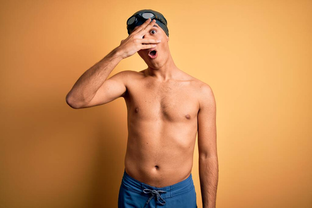 Young handsome man shirtless wearing swimsuit and swim cap over isolated yellow background peeking in shock covering face and eyes with hand, looking through fingers with embarrassed expression. - Zdjęcie, obraz