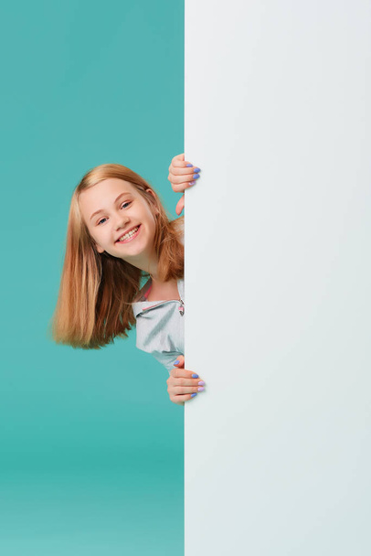 a laughing girl standing against a turquoise background holds an empty white Board in her hand and looks out from behind it. - Foto, Imagen