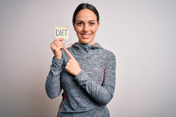 Joven mujer fitness usando ropa de entrenamiento deportivo sosteniendo papel con palabra de dieta muy feliz señalando con la mano y el dedo
 - Foto, imagen
