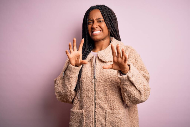 Junge afrikanisch-amerikanische Frau in modischem Wintermantel über rosa isoliertem Hintergrund angewiderte Miene, ungehalten und ängstlich dabei Ekel Gesicht, weil Abneigung Reaktion. Mit erhobenen Händen - Foto, Bild