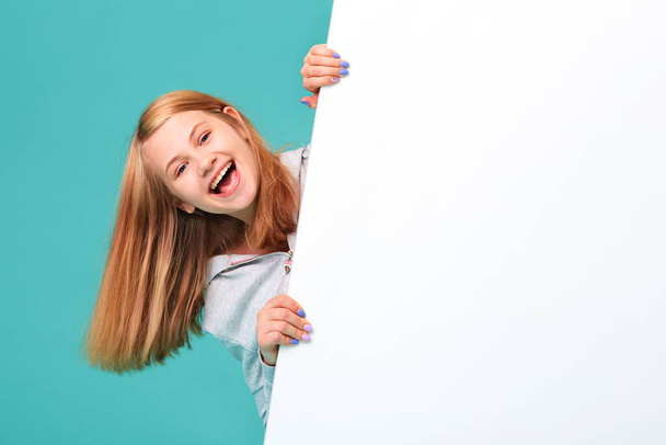 a laughing girl standing against a turquoise background holds an empty white Board in her hand and looks out from behind it. - Foto, Bild