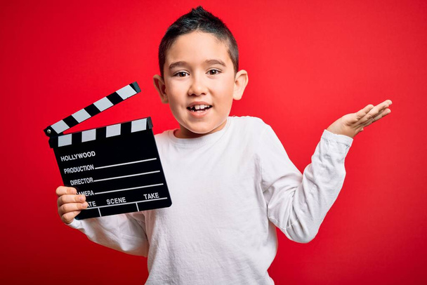 Nuori poika lapsi kuvaamassa video tilalla elokuvaohjaaja liitutaulu yli eristetty punainen tausta erittäin onnellinen ja innoissaan, voittaja ilme juhlii voittoa huutaen iso hymy ja nosti kädet
 - Valokuva, kuva