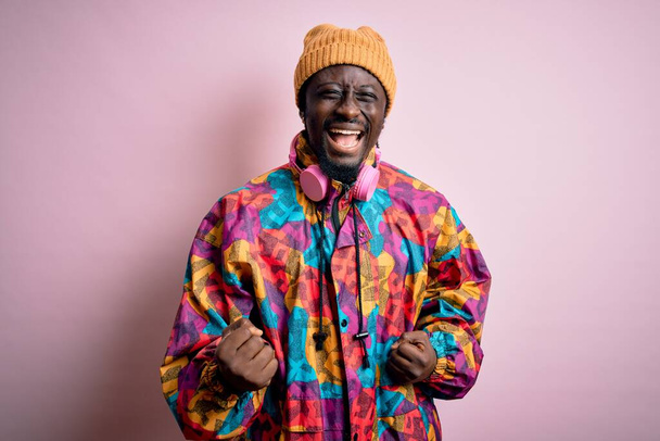 Jonge knappe Afro-Amerikaanse man draagt kleurrijke jas en pet over roze achtergrond erg blij en enthousiast doen winnaar gebaar met armen opgeheven, glimlachen en schreeuwen voor succes. Feestconcept. - Foto, afbeelding