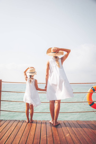 Suntanned woman and girl in white dresses enjoy sea view at the wooden pier - Photo, image