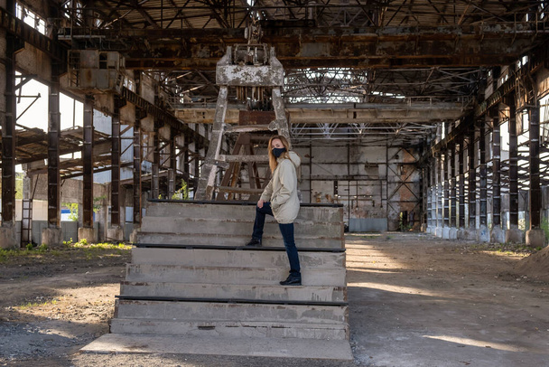 Een jong meisje met een zwart masker in het verwoeste gebouw van de oude fabriek. Gevolgen van de Apocalyps, epidemie, vernietiging. - Foto, afbeelding