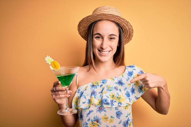 Młoda piękna turystka na wakacjach w letniej czapce pijąca napój koktajlowy z zaskakującą twarzą wskazującą palcem na siebie - Zdjęcie, obraz