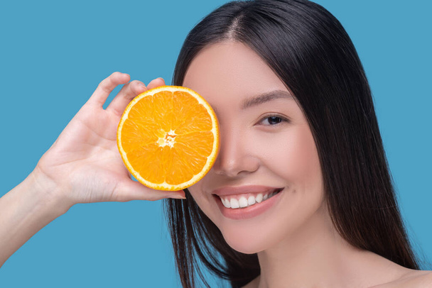 Smiling cute young woman holding a slice of orange and feeling pleased - Foto, afbeelding