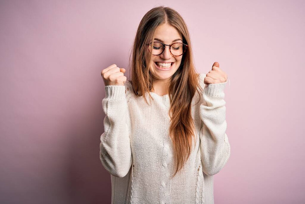 Young beautiful redhead woman wearing casual sweater and glasses over pink background excited for success with arms raised and eyes closed celebrating victory smiling. Winner concept. - Photo, image
