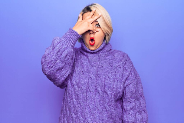 Beautiful blonde plus size woman wearing casual turtleneck sweater over purple background peeking in shock covering face and eyes with hand, looking through fingers with embarrassed expression. - Photo, Image