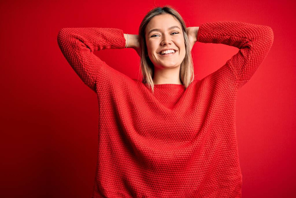 Giovane bella donna bionda indossa maglione casual su sfondo rosso isolato rilassante e stretching, braccia e mani dietro la testa e il collo sorridente felice
 - Foto, immagini