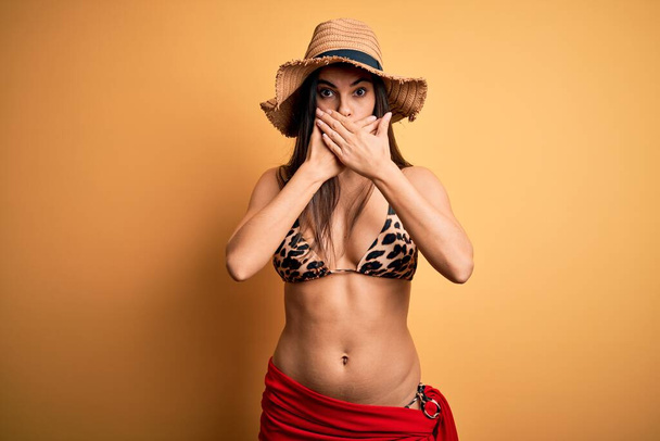 Mladá krásná brunetka žena na dovolené v plavkách a letním klobouku šokované zakrytí úst s rukama za omyl. Tajný koncept. - Fotografie, Obrázek