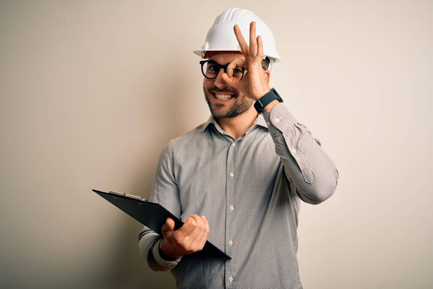 Νεαρός επιθεωρητής αρχιτέκτονα άνθρωπος φορώντας κράνος ασφαλείας οικοδόμος και χρησιμοποιώντας πρόχειρο με χαρούμενο πρόσωπο χαμογελώντας κάνει ok σημάδι με το χέρι στο μάτι κοιτάζοντας μέσα από τα δάχτυλα - Φωτογραφία, εικόνα