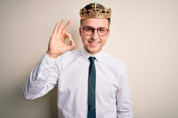 Young handsome caucasian business man wearing golden crown over isolated background smiling positive doing ok sign with hand and fingers. Successful expression. - Фото, изображение