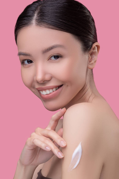 Pretty asian girl applying cream on her body and looking pleased - Valokuva, kuva