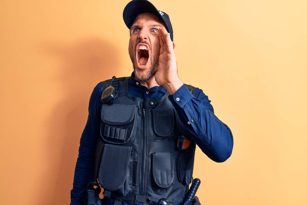 Young handsome policeman wearing police uniform and bulletprof over yellow background shouting and screaming loud to side with hand on mouth. Communication concept. - Photo, Image