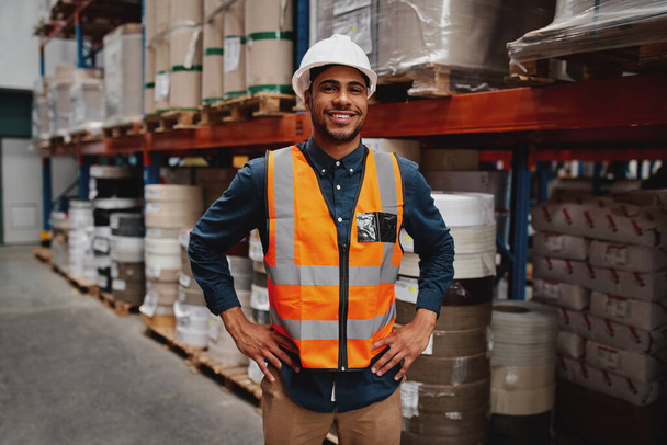 Portrait de gestionnaire heureux debout avec ses mains sur la taille portant un casque blanc et gilet de sécurité dans l'entrepôt avec des étagères d'inventaire en arrière-plan
 - Photo, image