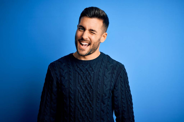Young handsome man wearing casual sweater standing over isolated blue background winking looking at the camera with sexy expression, cheerful and happy face. - Photo, image