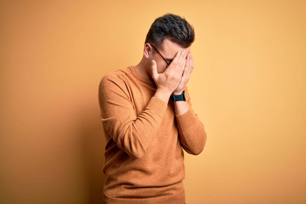 Young handsome caucasian man wearing glasses and casual winter sweater over yellow background with sad expression covering face with hands while crying. Depression concept. - Fotoğraf, Görsel