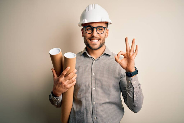 Młody architekt noszący kask wykonawcy trzymający papierowy plan projektu na odosobnionym tle robi znak ok palcami, doskonały symbol - Zdjęcie, obraz