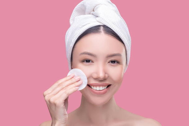 Pretty asian girl in a white headscarf cleaning her face with a sponge and feeling happy - Foto, afbeelding