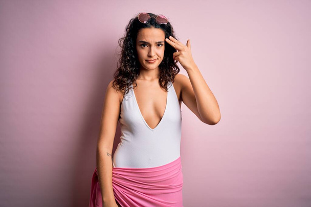 Beautiful woman with curly hair on vacation wearing white swimsuit over pink background Shooting and killing oneself pointing hand and fingers to head like gun, suicide gesture. - Photo, Image