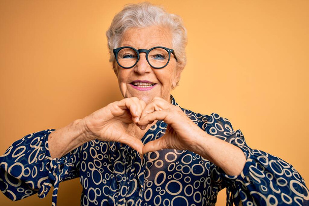 Senior beautiful grey-haired woman wearing casual shirt and glasses over yellow background smiling in love showing heart symbol and shape with hands. Romantic concept. - Photo, Image