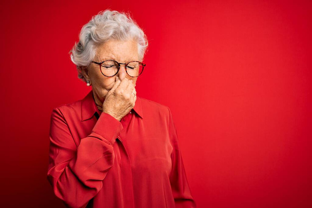 Starší krásná šedovlasá žena v neformální košili a brýlích na červeném pozadí ucítila něco páchnoucího a odporného, nesnesitelného, zadržující dech s prsty na nose. Špatný zápach - Fotografie, Obrázek