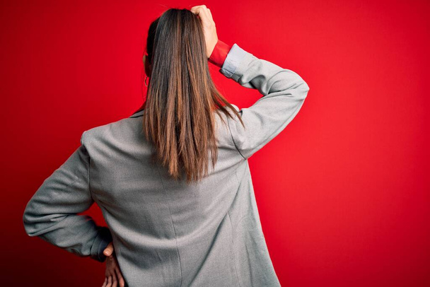 Young beautiful brunette businesswoman wearing jacket and glasses over red background Backwards thinking about doubt with hand on head - Valokuva, kuva