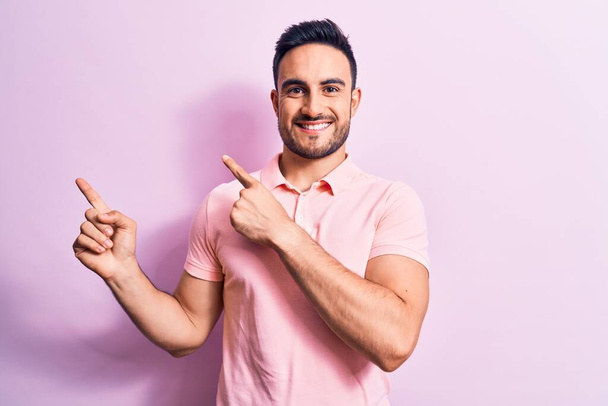 Young handsome man with beard wearing casual polo standing over pink background smiling and looking at the camera pointing with two hands and fingers to the side. - Fotó, kép