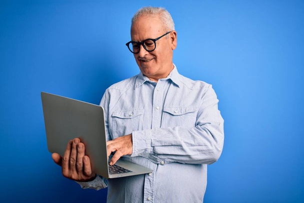 Красивый седой бизнесмен средних лет в очках, работающий с ноутбуком, со счастливым лицом, стоящим и улыбающимся с уверенной улыбкой, показывая зубы - Фото, изображение