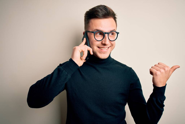Young handsome caucasian man having a conversation talking on smartphone pointing and showing with thumb up to the side with happy face smiling - Zdjęcie, obraz