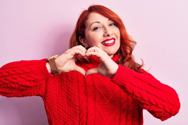 Young beautiful redhead woman wearing red casual turtleneck sweater over pink background smiling in love doing heart symbol shape with hands. Romantic concept. - Photo, Image