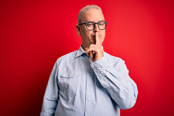 Middelbare leeftijd knappe hijgende man draagt casual gestreepte shirt en bril over rode achtergrond vragen om stil te zijn met de vinger op de lippen. Stilte en geheim concept. - Foto, afbeelding
