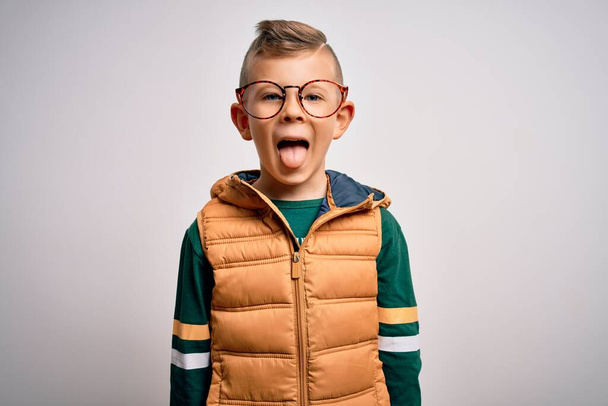 Ein kleines kaukasisches Kind mit blauen Augen, Wintermantel und smarter Brille, die die Zunge mit lustigem Gesichtsausdruck herausstreckt. Emotionales Konzept. - Foto, Bild