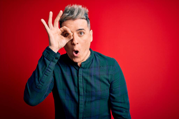 Young handsome modern man wearing elegant green shirt over red isolated background doing ok gesture shocked with surprised face, eye looking through fingers. Unbelieving expression. - Photo, Image