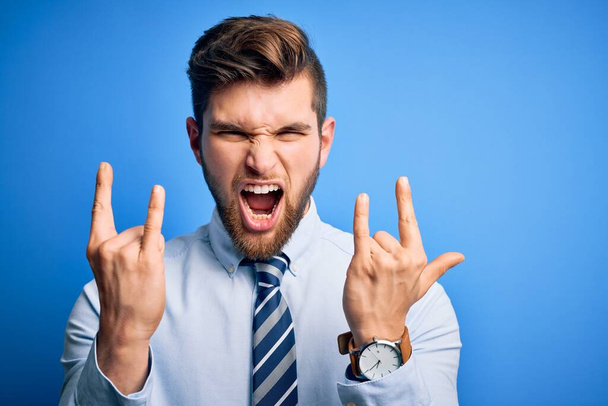 Young blond businessman with beard and blue eyes wearing elegant shirt and tie standing shouting with crazy expression doing rock symbol with hands up. Music star. Heavy concept. - Фото, изображение