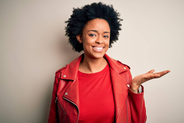Junge schöne afroamerikanische Afro-Frau mit lockigem Haar trägt lässig rote Jacke lächelnd fröhlich präsentiert und zeigt mit der Handfläche in die Kamera. - Foto, Bild