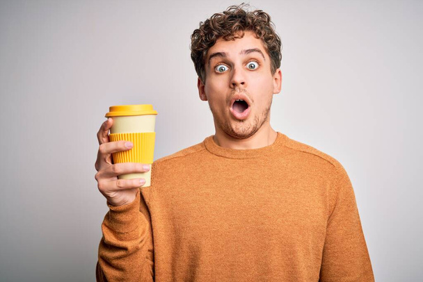 Jeune homme blond aux cheveux bouclés buvant une tasse de café debout sur fond blanc effrayé en état de choc avec un visage surprise, effrayé et excité par l'expression de la peur
 - Photo, image