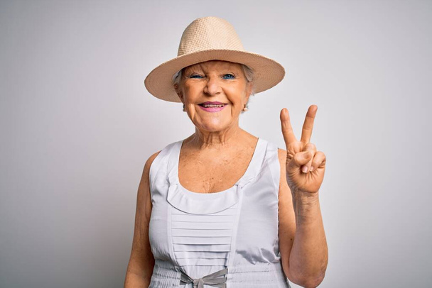 Senior mooie grijs-harige vrouw op vakantie in casual zomerjurk en hoed glimlachend met gelukkig gezicht knipogen naar de camera doen overwinning teken. Nummer twee. - Foto, afbeelding