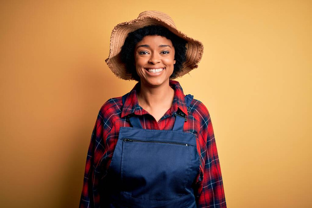 Young African American afro farmer woman with curly hair wearing apron and hat with a happy and cool smile on face. Lucky person. - Photo, image