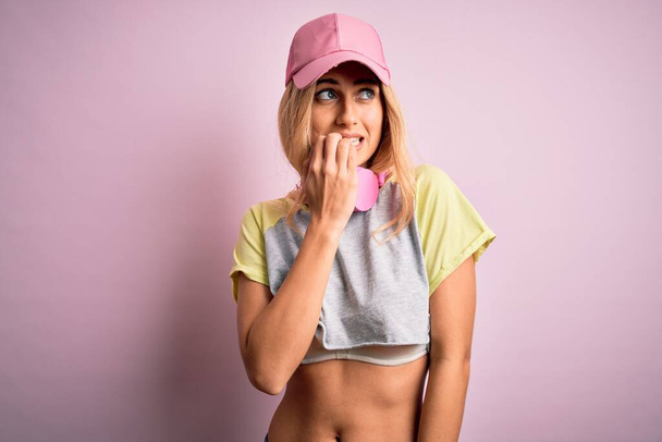 Młoda piękna blondynka sportowiec robi sport za pomocą słuchawek na różowym tle patrząc zestresowany i nerwowy z rękami na ustach obgryzając paznokcie. Problem lękowy. - Zdjęcie, obraz