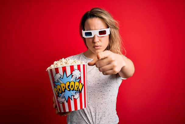 Młoda piękna blondynka ogląda film przy użyciu okularów 3D jedząc popcorn snack wskazując palcem na aparat i do Ciebie, znak ręczny, pozytywny i pewny gest z przodu - Zdjęcie, obraz