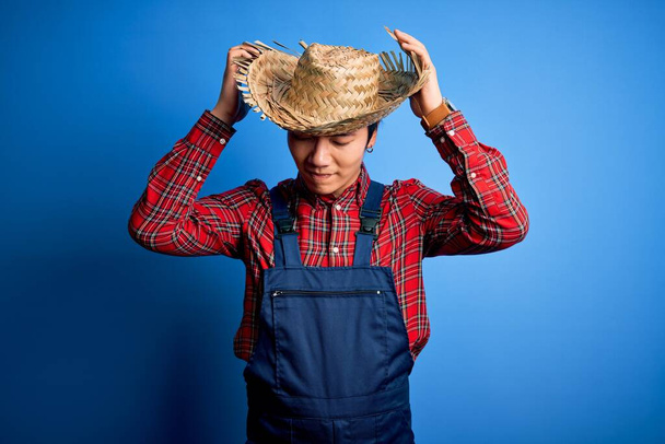 Joven granjero chino guapo que usa delantal y sombrero de paja sobre fondo azul que sufre de dolor de cabeza desesperado y estresado porque el dolor y la migraña. Manos en la cabeza
. - Foto, imagen